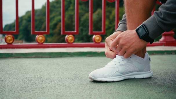 Vista recortada de deportista con smartwatch atando cordones de zapatilla en puente - Imágenes, Vídeo
