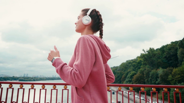 Zijaanzicht van gelukkige sportvrouw met koptelefoon en smartwatch draait op de brug - Video