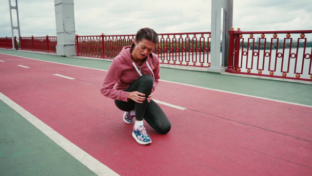 Kobieta biegacz z smartwatch dotykając bolącej nogi podczas siedzenia na ścieżce mostu - Materiał filmowy, wideo