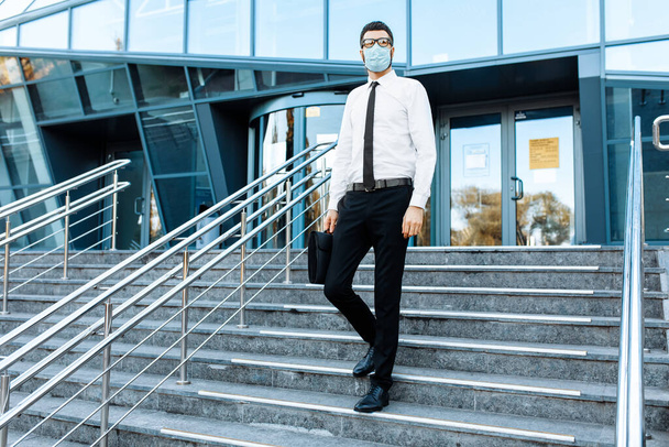 young businessman in business clothes, a man in a medical protective mask on his face, leaves the office building after a working day - Fotoğraf, Görsel