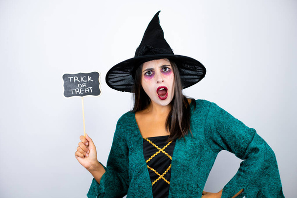 Mulher vestindo traje de bruxa sobre fundo branco isolado surpreendido e segurando quadro negro com truque ou mensagem palavra tratar - Foto, Imagem