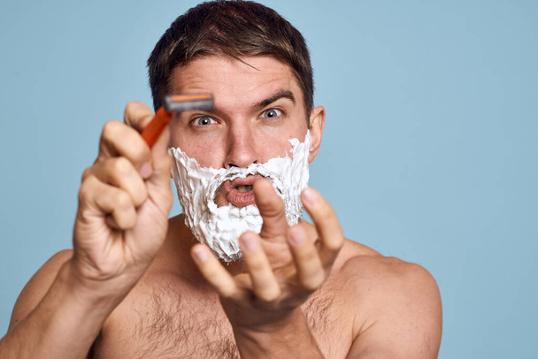 Man met scheermes en wit schuim op zijn gezicht blauwe achtergrond naakt romp baden procedures - Foto, afbeelding