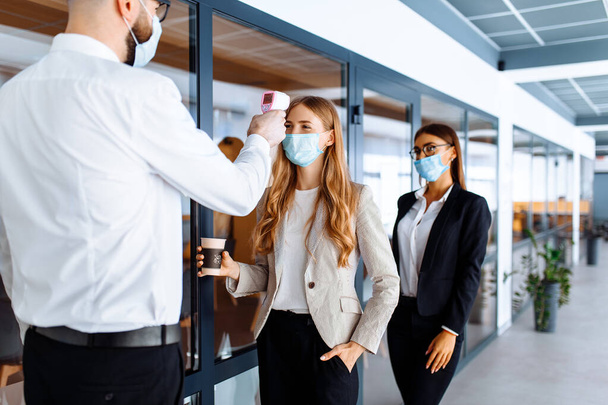 man with a non-contact thermometer measures the temperature of employees in protective masks at the entrance to a modern office - Zdjęcie, obraz