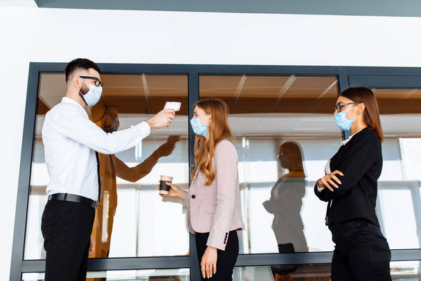Mann mit berührungslosem Thermometer misst die Temperatur von Mitarbeitern in Schutzmasken am Eingang eines modernen Büros - Foto, Bild