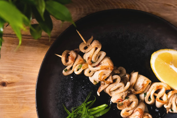 Fried squid bbq on skewers with lemon lies on a dark plate. Restaurant food and service concept - Foto, Imagem