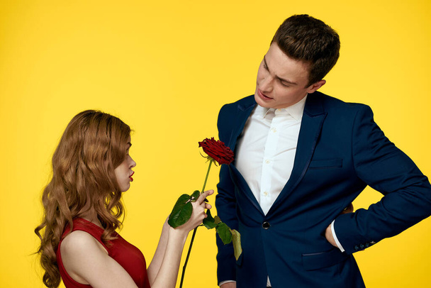 Een romantische man knuffelt een vrouw in een rode jurk met een roos in zijn hand op een gele achtergrond - Foto, afbeelding