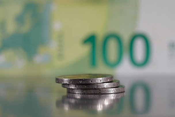 Una pequeña pila de tres monedas se ha apilado sobre una superficie gris y plana y es visible frente a un borroso billete de 100 euros.. - Foto, imagen