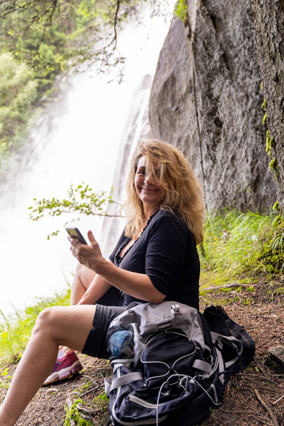 smiling woman on vacation sitting on a stone in front of a mountain waterfall - Fotó, kép