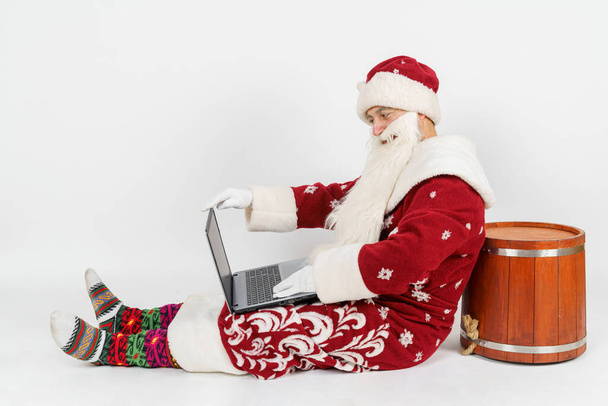Christmas and New Years concept. Santa Claus is sitting on the floor and working at a laptop. Isolated background. - Фото, изображение