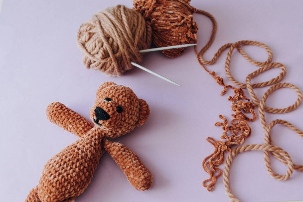 Hobby and stuffed toys concept. Place for text and add. Brown knitting yarn. The knitting needles are stuck in the yarn. Knitted brown soft bear on a lavender background.  - Photo, Image