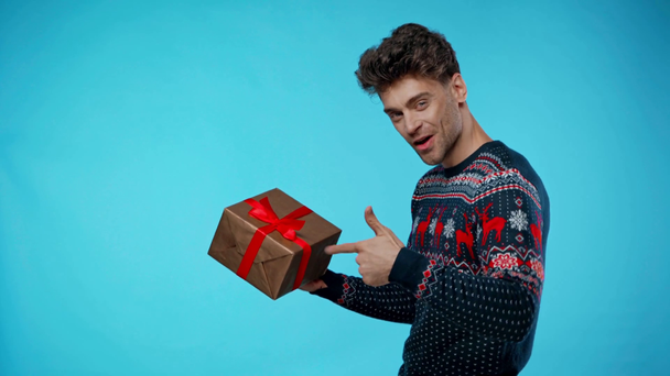 Fröhlicher junger erwachsener Mann zeigt mit dem Finger auf Geschenkbox auf blau - Filmmaterial, Video
