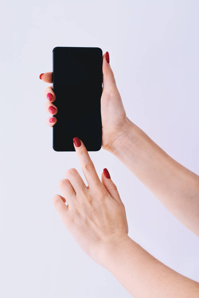 Elegant female hands with red nails holding a smart phone and pointing the touchscreen. Closeup on light grey background - Photo, Image