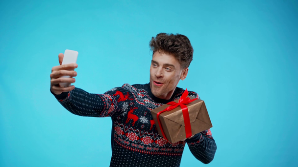 Ενθουσιασμένος νεαρός άνδρας που παίρνει selfie ενώ κρατά κουτί δώρου στο μπλε - Πλάνα, βίντεο