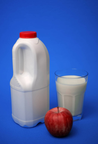 Airtight one gallon milk jug with a red cap, full glass milk and red apple on a blue background - Photo, Image