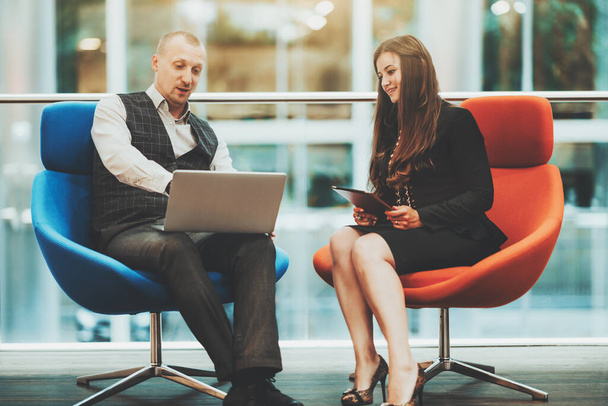 Two business partners are sitting on armchairs or blue and orange color: a man entrepreneur with a laptop and his female colleague a businesswoman with a tablet pc in hands, they are having a talk - Foto, afbeelding