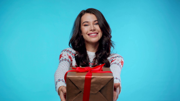Happy young adult woman showing gift box, while looking at camera on blue - Footage, Video