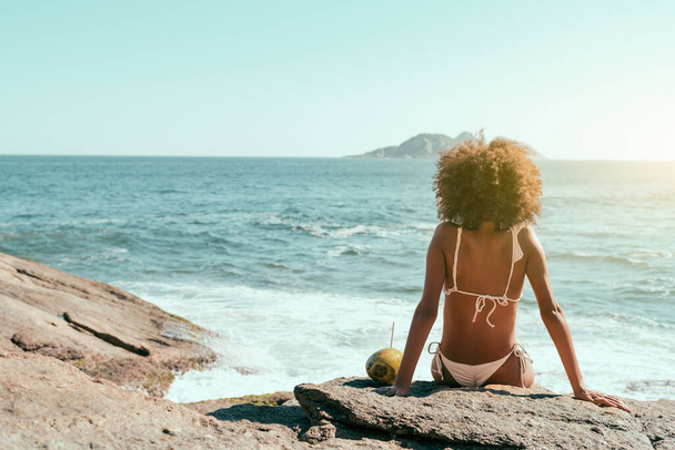 View from behind of a young African-American female with curly hair, in a swimsuit sitting on a rock near the beach of a resort and enjoying the sun, observing seascape, relaxing; a coconut near her - Foto, Bild