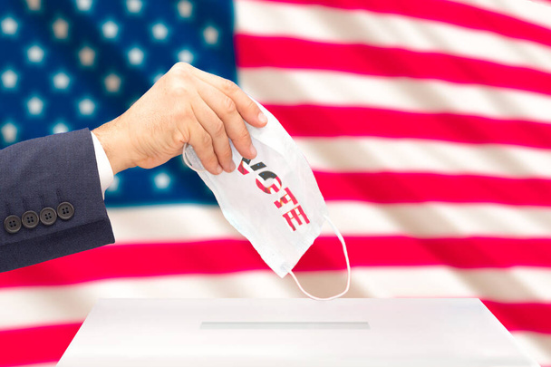 Το χέρι ενός ανθρώπου που ψηφίζει στις προεδρικές εκλογές των ΗΠΑ με μια ιατρική μάσκα τη σημαία των ΗΠΑ πίσω από αυτό. Επιλεκτική εστίαση - Φωτογραφία, εικόνα