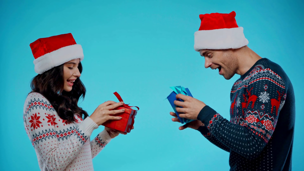 Boční pohled na šťastný pár v Santa Hats dávat dárky k sobě na modré - Záběry, video