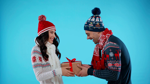 Side view of happy couple in knitted hats and scarfs holding gift box on blue - Footage, Video