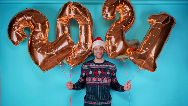 Glücklicher junger erwachsener Mann mit Weihnachtsmütze und goldenen Luftballons mit Zahlen auf blauem Grund - Filmmaterial, Video
