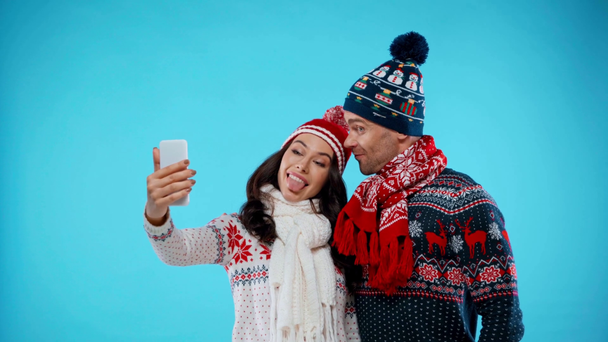 gelukkig paar in hoeden en sjaals het nemen van selfie en tonen grimaces op blauw - Video