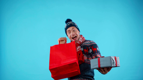 Heyecanlı bir adam elinde hediye kutusu ve mavi üzerinde kırmızı kağıt torba tutuyor. - Video, Çekim
