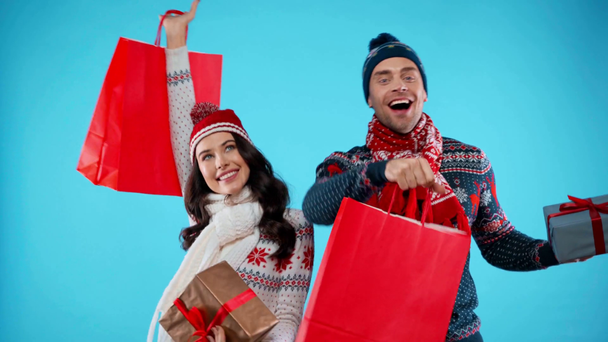 Smiling couple showing red paper bags, while holding gift boxes on blue - Footage, Video