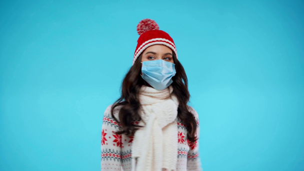 Νεαρή ενήλικη γυναίκα με πλεκτό μαντήλι και καπέλο που χορεύει, φορώντας ιατρική μάσκα στο μπλε - Πλάνα, βίντεο