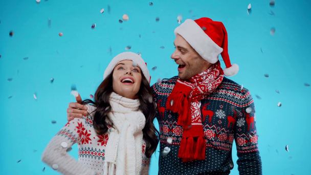 jong paar in santa hoeden omarmen, terwijl confetti vallen op blauw - Video