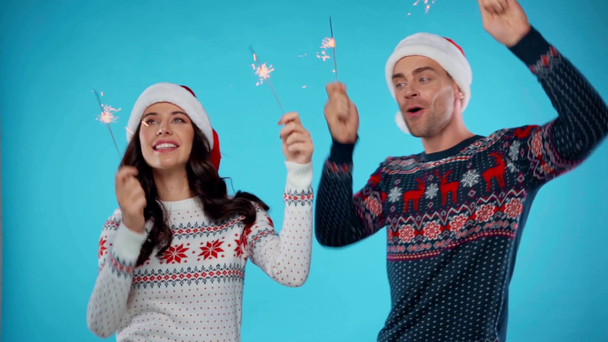 Heureux jeune couple en chapeaux santa tenant des étincelles et dansant sur bleu - Séquence, vidéo
