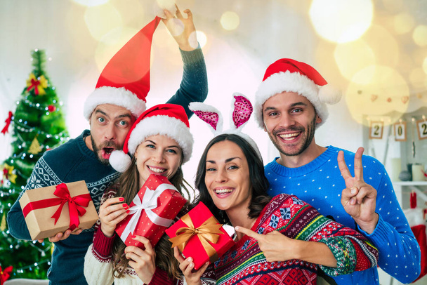 Happy group of excited beautiful friends in Santa hats are having fun while partying at home on the Christmas tree background - 写真・画像