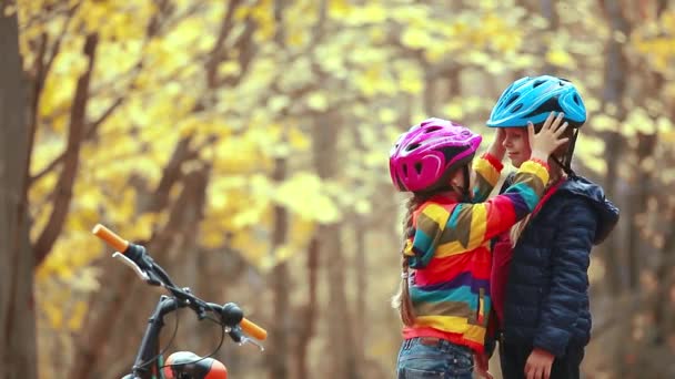 Младшая сестра помогает старшей девушке надеть защитный шлем перед велоспортом в осенний день на природе и дать друг другу пять. Безопасность, спорт, отдых - Кадры, видео