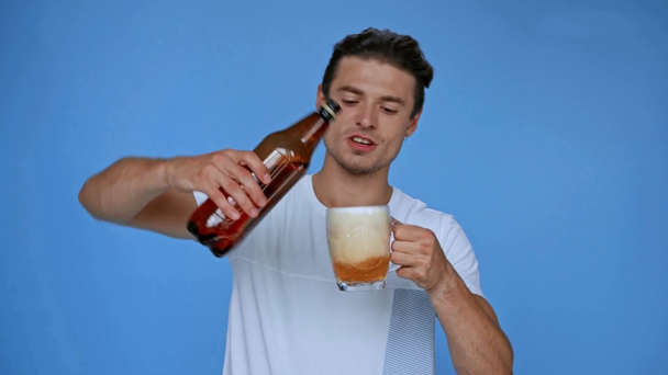 hombre feliz en camiseta blanca vertiendo cerveza en vidrio aislado en azul - Metraje, vídeo