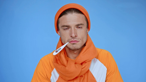 homem doente em chapéu e lenço medição temperatura isolada em azul - Filmagem, Vídeo