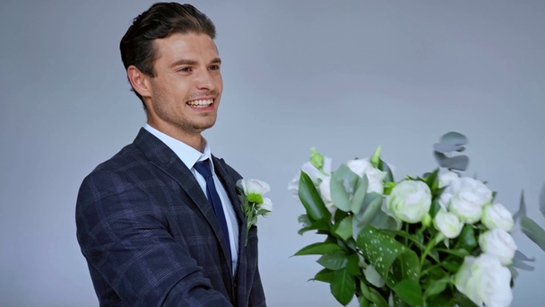 novio feliz hablando y dando flores a la novia aislado en gris  - Metraje, vídeo