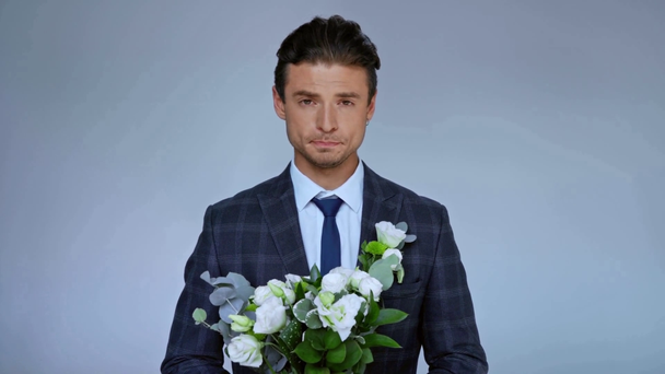 γαμπρός με κοστούμι κρατώντας λουλούδια και χαμογελώντας απομονωμένος σε γκρι - Πλάνα, βίντεο