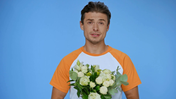 hombre feliz en camiseta sosteniendo flores mientras habla aislado en azul - Imágenes, Vídeo