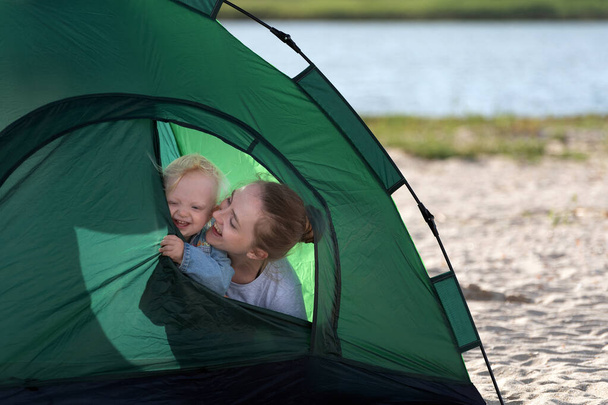 Улыбающиеся мама и ребенок выглядывают из туристической палатки. Палатка на песчаном пляже. Кемпинг. - Фото, изображение