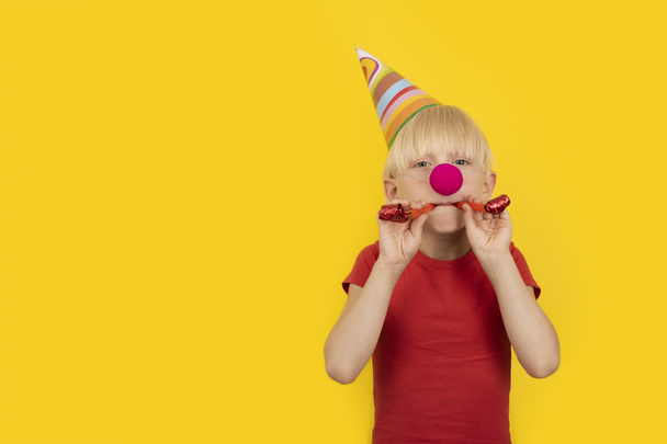 Garçon avec chapeau de fête et nez de clown rouge tenant sifflet. Portrait sur fond jaune. Matinée de vacances. - Photo, image