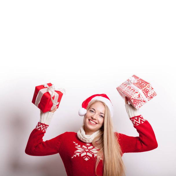 smiling blond millennial lady with santa hat and knitted sweater holds two christmas presents in her hands - Photo, image