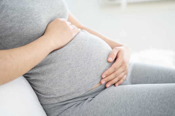Nahaufnahme einer schwangeren Frau, die zu Hause auf dem Bett sitzt und ihren Bauch berührt. Selektiver Fokus. - Foto, Bild