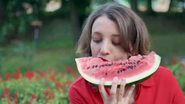 Schöne Frau in rotem Hemd mit kurzen Haaren sitzt draußen auf rotem Blumenhintergrund und isst ein Stück Wassermelone. Picknick in der Stadt. - Filmmaterial, Video
