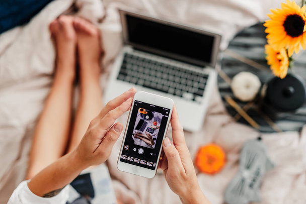 Woman is taking a photo using a phone. Overhead picture. Snapping an image. Bedroom morning light, comfortable cozy Autumn mood. Laptop, pumpkin, coffee. Fall vibes. Cozy lazy morning. Social media - Fotó, kép