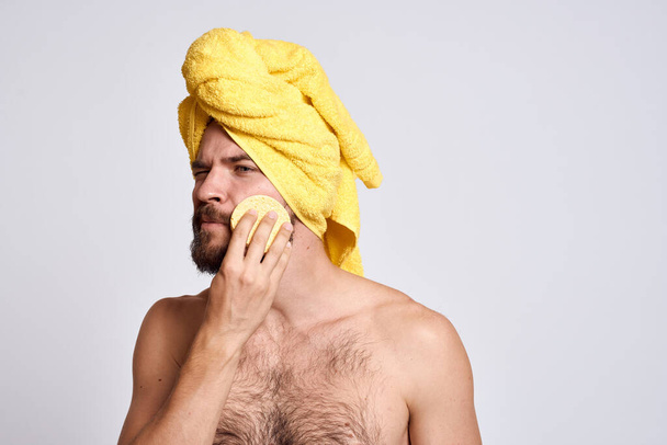 Mann mit einem gelben Handtuch auf dem Kopf nackte Schultern Schwamm saubere Haut Gesichtspflege Licht Hintergrund - Foto, Bild
