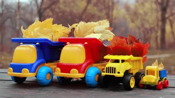 Cztery małe zabawki ciężarówka jest załadowana żółtymi spadającymi liśćmi. Samochód stoi na drewnianej powierzchni na tle rozmytego jesiennego parku. Czyszczenie i usuwanie opadłych liści. Roboty sezonowe - Materiał filmowy, wideo