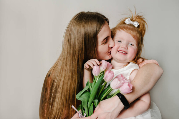 The mother kissing little girl on light background indoor. Portrait of mom with flowers and daughter sitting in home. The concept of happy family holiday. woman's day. Mother's Day. - Photo, image