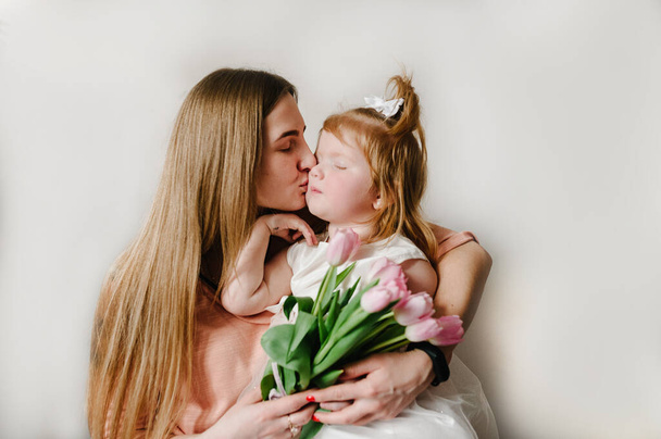 La mère embrassant la petite fille sur fond clair à l'intérieur. Portrait de maman avec des fleurs et fille assise à la maison. Le concept de vacances en famille heureuse. le jour de la femme. Fête des mères. - Photo, image