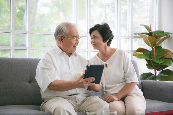 Seniorenpaar lächelt und schaut auf dem Sofa auf das gleiche Tablet. - Foto, Bild