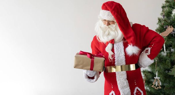 Święty Mikołaj stoi obok choinki, trzymając pudełko z prezentem i patrząc na nie. przestrzeń kopiowania - Zdjęcie, obraz
